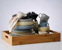 Et sæt keramikservice i afdæmpede nuancer af blå, grå og beige, herunder tallerkener, skåle og krus, pænt stablet i en rektangulær bakke af træ. Sættet har en glat, blank overflade og et minimalistisk design. En cremefarvet stofserviet og en klase kunstige sorte druer ligger ovenpå som dekoration.
