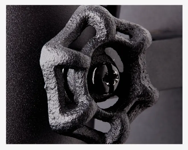 Nærbillede af et sort, støbejernshåndtag med hjulform og mat overflade. Håndtaget har en ru, industriel struktur og en blank sort skrue eller bolt i midten. Baggrunden er mørk og uskarp, hvilket fremhæver håndtagets detaljer.