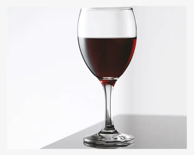 Et klart vinglas med stilk, fyldt halvt op med mørkerød væske. Glasset har en rund skål og en solid fod, og det står på en reflekterende overflade med en lys, ensfarvet baggrund.