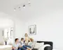 Lyst, minimalistisk stue med sort hjørnesofa, hvide puder, indrammet vægkunst og en loftmonteret spotlight-skinne; en familie på fire sidder og leger sammen i sofaen.