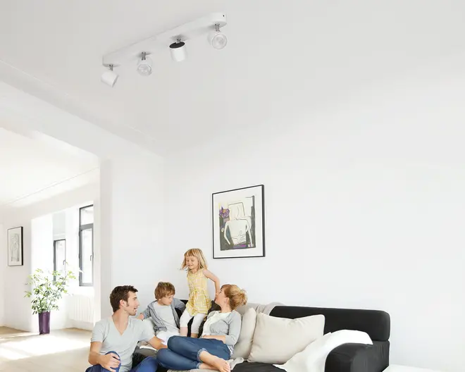 Lyst, minimalistisk stue med sort hjørnesofa, hvide puder, indrammet vægkunst og en loftmonteret spotlight-skinne; en familie på fire sidder og leger sammen i sofaen.