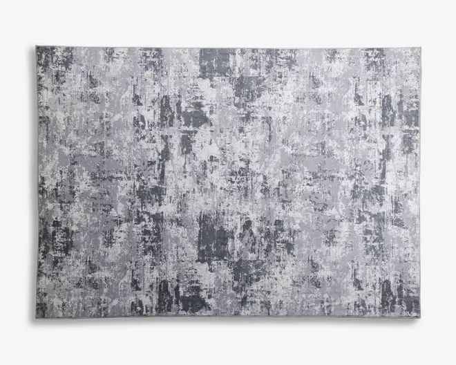Rektangulært tæppe med et moderne abstrakt mønster i forskellige nuancer af grå og hvid. Designet har et slidt, tekstureret udseende med uregelmæssige felter og penselstrøg-lignende detaljer, hvilket giver det et moderne og alsidigt udtryk. Overfladen ser blød og flad ud, velegnet til mange forskellige indretningsstile.