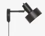 Mat sort vægmonteret lampe med et cylindrisk justerbart arm og en bred, vinklet lampeskærm. Lampen har en synlig ledning, der hænger ned fra basen, og skærmen kan vippes for at rette lyset. Designet er moderne og minimalistisk, velegnet til fokuseret belysning.