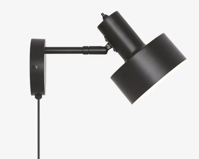 Mat sort vægmonteret lampe med et cylindrisk justerbart arm og en bred, vinklet lampeskærm. Lampen har en synlig ledning, der hænger ned fra basen, og skærmen kan vippes for at rette lyset. Designet er moderne og minimalistisk, velegnet til fokuseret belysning.