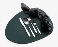 Et sæt med fire bestikdele i rustfrit stål, herunder en kniv, en spiseske, en gaffel og en teske, alle med et slankt, moderne design og glatte håndtag. Bestikket er pænt arrangeret på en mørk, uregelmæssigt formet dækkeserviet. Der ses også en mørk stofserviet bundet i en knude samt en klase sorte druer, hvilket giver et elegant udtryk.