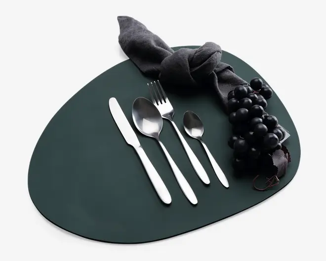 Et sæt med fire bestikdele i rustfrit stål, herunder en kniv, en spiseske, en gaffel og en teske, alle med et slankt, moderne design og glatte håndtag. Bestikket er pænt arrangeret på en mørk, uregelmæssigt formet dækkeserviet. Der ses også en mørk stofserviet bundet i en knude samt en klase sorte druer, hvilket giver et elegant udtryk.