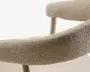 Nærbillede af en beige tekstureret polstret stol med buet armlæn og blød ryg, båret af slanke lyse træben.