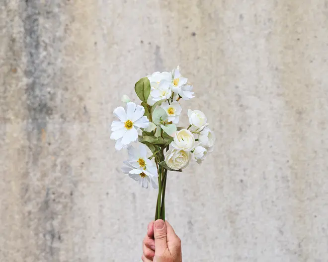 En hånd holder en lille buket kunstige hvide blomster med gule midter og grønne blade. Buketten består af en blanding af margueritlignende og roselignende blomster med realistiske stilke og løv. Baggrunden er en ensfarvet, lys og let tekstureret væg.