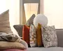 En samling dekorative puder i blandede ტექստurer og mønstre, herunder striber, blomster, ensfarvede og geometriske betræk i beige, orange, sennep, grå og hvid, arrangeret på en lysegrå sofa med en høj gulvlampe bagved.