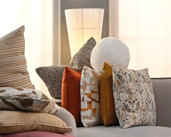 En samling dekorative puder i blandede ტექստurer og mønstre, herunder striber, blomster, ensfarvede og geometriske betræk i beige, orange, sennep, grå og hvid, arrangeret på en lysegrå sofa med en høj gulvlampe bagved.