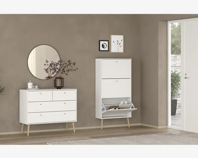 Billedet viser to moderne møbler med minimalistisk design, begge i mat hvid finish med lyse træben og små gyldne greb. Til venstre står en kommode med tre store skuffer og to mindre øverste skuffer, hvilket giver god opbevaringsplads. Til højre ses et højt, slankt skab med to rum; det nederste rum er åbent og viser organiseret skoopbevaring med skrå hylder. Møblerne står mod en taupefarvet væg, suppleret af et rundt spejl, en dekorativ vase med grene og indrammet vægkunst over skabet. Udtrykket er rent, moderne og funktionelt.