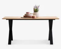 Rektangulært bord med en lys træbordplade og sorte, skråtstillede metalben. Bordpladen er pyntet med en stak lyserøde keramiktallerkener og skåle, en matchende kop, en rund fletbrik, en lille lyserød skål og en hvid vase med tekstur, der indeholder lilla og grønne kunstige blomster.