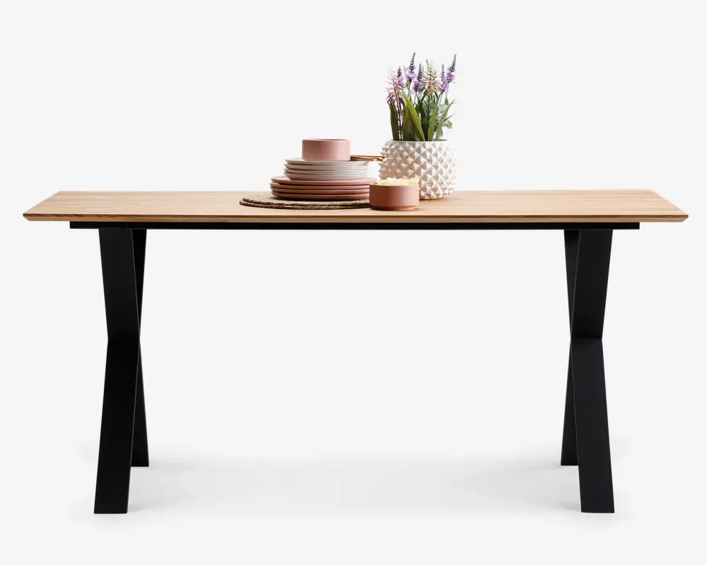 Rektangulært bord med en lys træbordplade og sorte, skråtstillede metalben. Bordpladen er pyntet med en stak lyserøde keramiktallerkener og skåle, en matchende kop, en rund fletbrik, en lille lyserød skål og en hvid vase med tekstur, der indeholder lilla og grønne kunstige blomster.