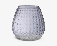 Klar glasbeholder med rund, bulet form og en tekstureret overflade med hævede cirkulære knopper arrangeret i et regelmæssigt mønster. Åbningen i toppen er lidt smallere end den bredeste del af kroppen. Glasset er gennemsigtigt og har et moderne, dekorativt udseende.