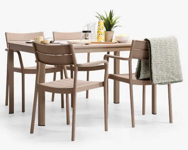 Et moderne spisesæt bestående af et rektangulært bord og seks matchende stole i en afdæmpet beige farve. Stolene har et enkelt design med lamel-ryg, og to af stolene har armlæn. Bordet er dækket med morgenmadsting, herunder en kop, skål, tallerkener, en karaffel, et glas og en dekorativ plante. Et gråt, tekstureret tæppe er lagt over ryggen på en af stolene, hvilket giver et hyggeligt præg.