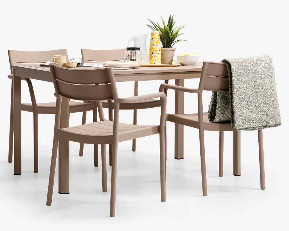 Et moderne spisesæt bestående af et rektangulært bord og seks matchende stole i en afdæmpet beige farve. Stolene har et enkelt design med lamel-ryg, og to af stolene har armlæn. Bordet er dækket med morgenmadsting, herunder en kop, skål, tallerkener, en karaffel, et glas og en dekorativ plante. Et gråt, tekstureret tæppe er lagt over ryggen på en af stolene, hvilket giver et hyggeligt præg.