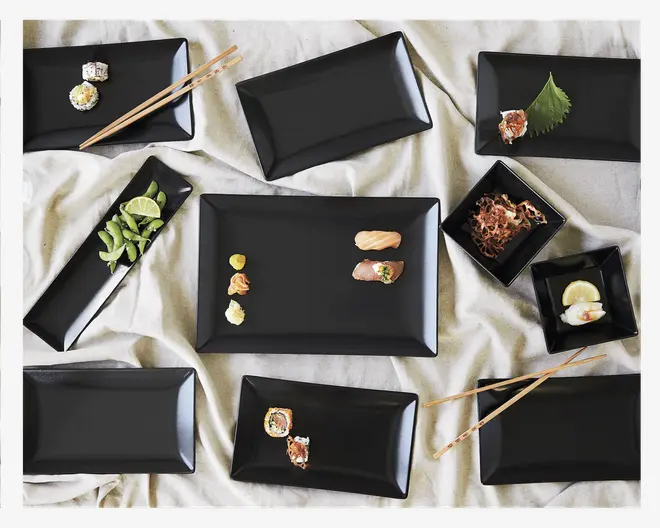 Et sæt elegante, matte sorte rektangulære og firkantede tallerkener arrangeret på et lyst stof. Tallerkenerne har let hævede kanter og et moderne, minimalistisk design. Nogle tallerkener har sushi, limebåde og pynt, mens andre er tomme og fremhæver den glatte, enkle overflade. Træspisepinde er også med, hvilket understreger sættets egnethed til servering af asiatisk mad.
