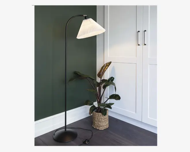 En høj, sort gulvlampe med buet hals og en plisseret, hvid lampeskærm. Lampen har en rund, stabil fod og en synlig fodkontakt på ledningen. Den står ved siden af en grøn væg og hvide skabe, med en fletkurv med en grøn plante i nærheden.