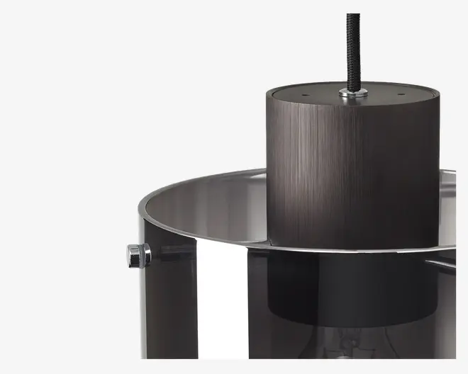 Nærbillede af en moderne pendellampe med et cylindrisk sort metalhus med børstet finish, ophængt i en ledning. Lampen har en halvgennemsigtig, mørktonet glasskærm med glat, blank overflade og kromdetaljer på siden. Designet er stilrent og moderne med rene linjer og et minimalistisk udtryk.
