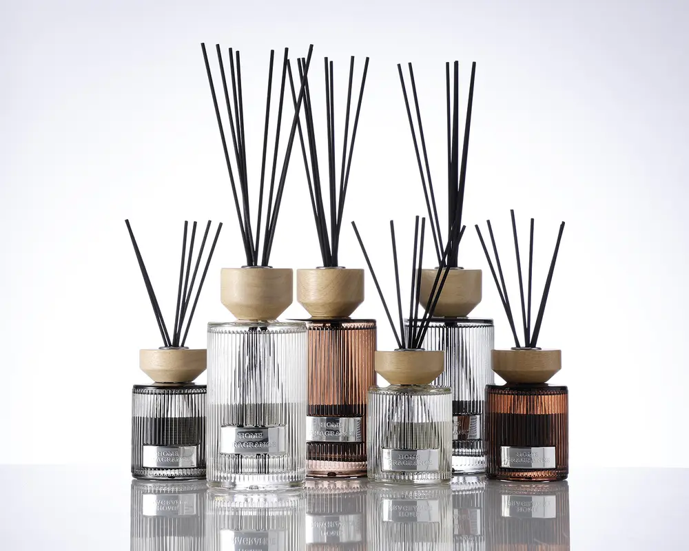 En samling dekorative glasbeholdere med lodrette rillede strukturer, hver med et lyst trælåg og fyldt med sorte duftpinde. Beholderne varierer i størrelse og farve, herunder klare, røggrå og ravfarvede nuancer. Hver beholder har en metallabel på forsiden, hvilket giver et eksklusivt og elegant udtryk. Det samlede design er moderne og stilfuldt, velegnet til boligindretning.