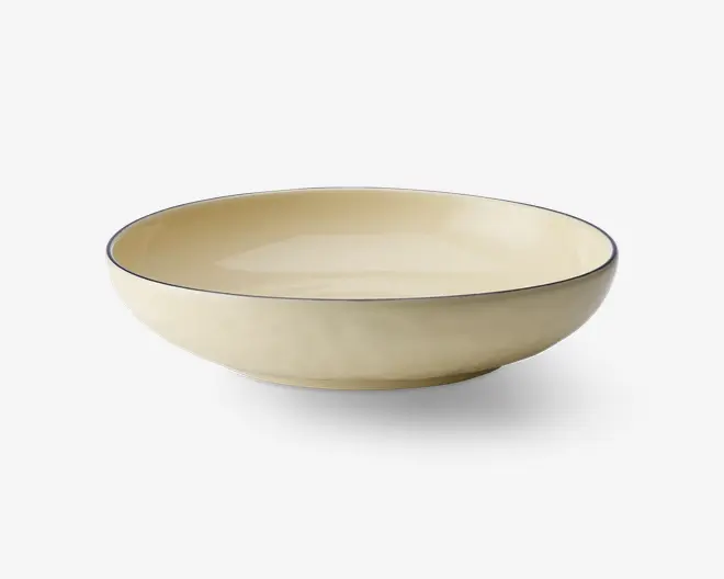 En rund, lav skål med en glat, blank cremefarvet overflade. Skålens kant er markeret med en tynd, mørk streg, som giver et diskret og elegant udtryk. Skålen har blødt skrånende sider og en flad bund, velegnet til servering af salater, pasta eller andre retter.