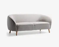 Et moderne, lysegråt polstret siddemøbel med buet ryglæn og armlæn, der flyder sammen med den samlede form. Sædet består af to bløde puder, og møblet står på fire let skrå, lyse træben, hvilket giver et stilrent og minimalistisk udtryk.