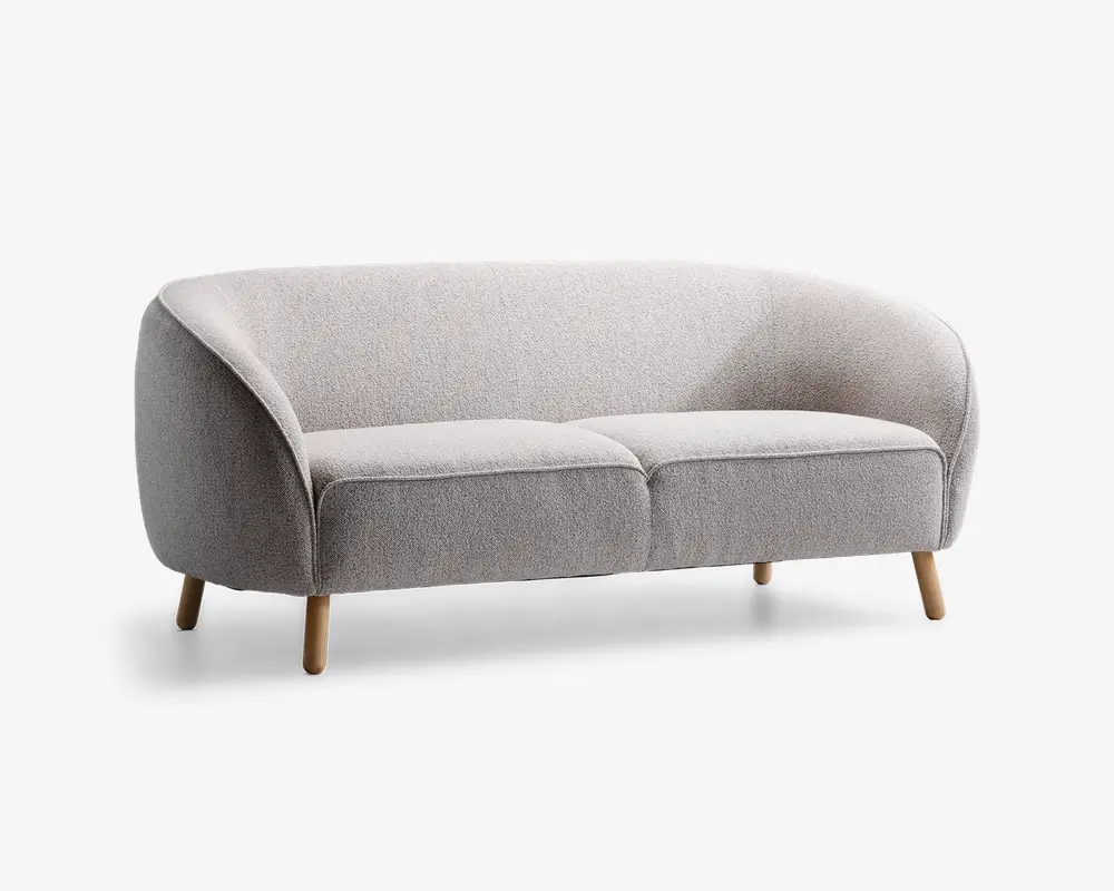 Et moderne, lysegråt polstret siddemøbel med buet ryglæn og armlæn, der flyder sammen med den samlede form. Sædet består af to bløde puder, og møblet står på fire let skrå, lyse træben, hvilket giver et stilrent og minimalistisk udtryk.