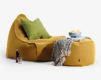 Et hyggeligt siddearrangement bestående af en loungestol i sennepsgul, tekstureret stof med en matchende puf. Stolen har en afslappet, blød form og er udstyret med en stor, blød grøn pude for ekstra komfort. Puffen er dækket af et grå- og hvidmønstret tæppe, og ovenpå står en blå skål med popcorn samt et magasin. Ved siden af stolen står en glasflaske med sodavand og et klart glas, hvilket giver et indbydende og afslappet indtryk.