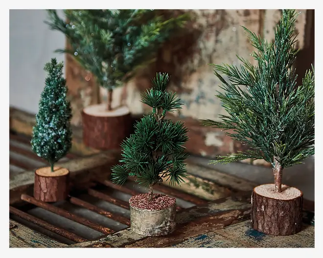 Sæt med fire kunstige miniature-grantræer, hver med forskellig form og type af grønne grene. Træerne står på små, runde baser, der ligner naturlige træskiver og giver et rustikt udtryk. Nogle af træerne har et lag kunstig sne på grenene, mens andre har et mere naturligt, bart udseende. Løvværket varierer fra tæt og fyldigt til mere spinkelt og spidst, hvilket giver forskellige teksturer. Disse dekorative elementer egner sig til bordpynt eller juleudsmykning.