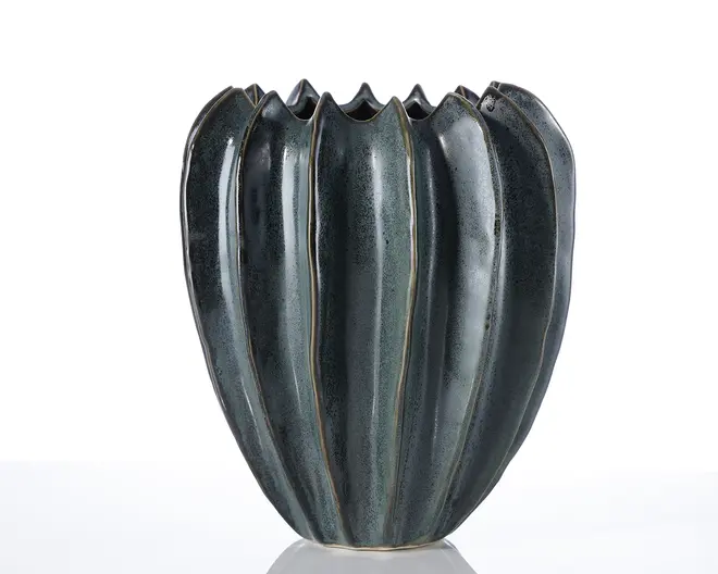 En keramisk vase med en mørk, blank overflade og et unikt, skulpturelt design. Vasen har lodrette riller, der danner en fløjet, organisk form, som minder om en kaktus eller blomsterknop. Kanten er ujævn og bølget, hvilket giver et kunstnerisk udtryk. Overfladen har diskrete farvevariationer med lysere nuancer langs rillernes kanter.