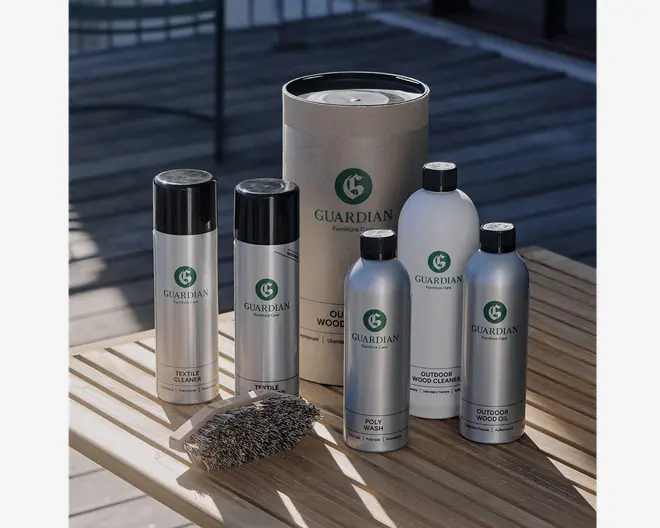 Et sæt rengørings- og vedligeholdelsesprodukter vist på et bord af træ, bestående af fem flasker og dåser mærket til forskellige formål såsom tekstilrens, polyvask, udendørs trærenser og udendørs træolie. Beholderne er primært sølvfarvede med grønne og sorte etiketter, og der er en rund paprørsemballage i baggrunden. Sættet inkluderer også en rengøringsbørste med stive børstehår og træskaft.