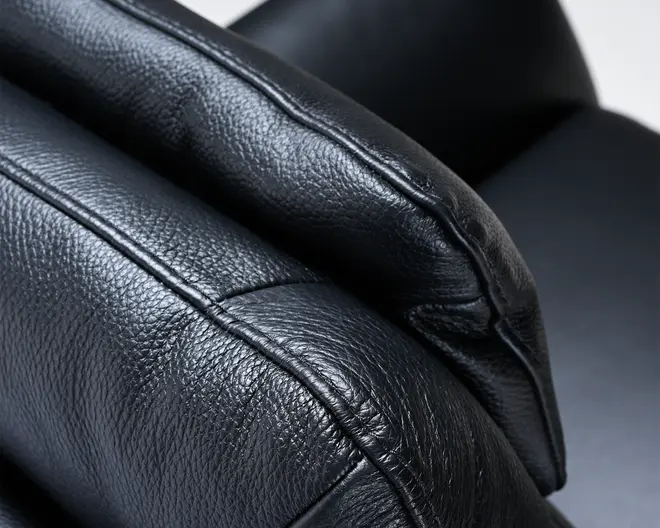 Nærbillede af en sort læderoverflade med synlig kornet tekstur og detaljerede syninger. Materialet ser blødt og polstret ud, hvilket indikerer komfort og støtte. Syningerne er pæne og forstærker kanterne, hvilket tyder på holdbarhed og god håndværksmæssig kvalitet.