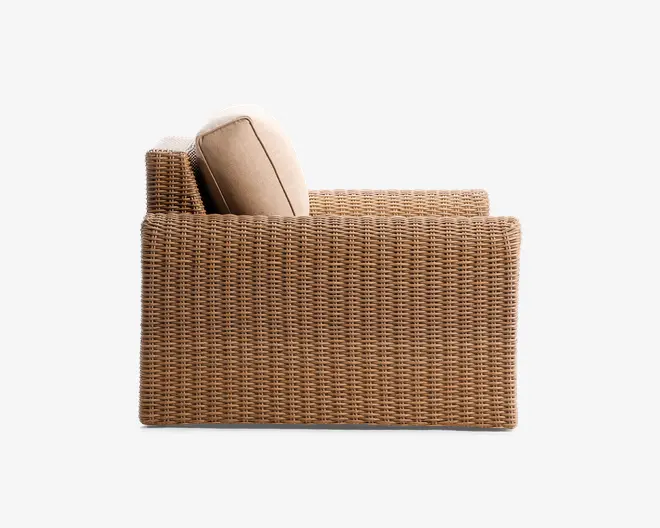 Sidebillede af et møbel med en tæt flettet rattan- eller fletramme i en naturlig lysebrun farve. Armlænet og ryglænet er lige og firkantede, hvilket giver et moderne udtryk. Der er en tyk, lys beige siddepude og en matchende rygpude, begge med synlige kanter for ekstra detalje og holdbarhed.