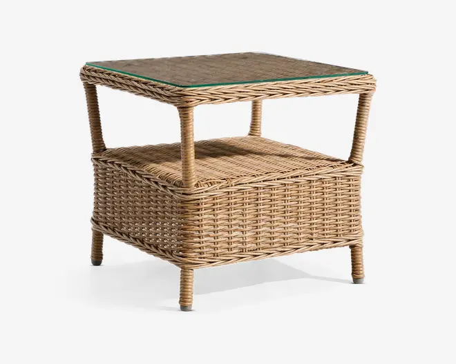 Firkantet sidebord fremstillet af naturfarvet flettet rattan med en glastop, der giver en jævn overflade. Bordet har en ekstra hylde nederst til opbevaring eller udstilling og står på fire solide, runde ben. Den flettede struktur giver et afslappet og let udtryk, der passer til både indendørs og udendørs brug.
