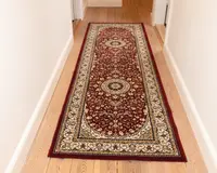 Lang, rektangulær tæppe med en dyb bordeaux baggrund og indviklede blomster-mønstre i creme, guld og sort. Tæppet har to store cirkulære medaljoner langs midten, omgivet af detaljerede vin- og blomstermotiver. En bred cremefarvet kant med dekorative mønstre omkranser tæppet, og kanterne er afsluttet med en ensfarvet bordeaux trim. Tæppet ligger på et lyst trægulv i en gang.