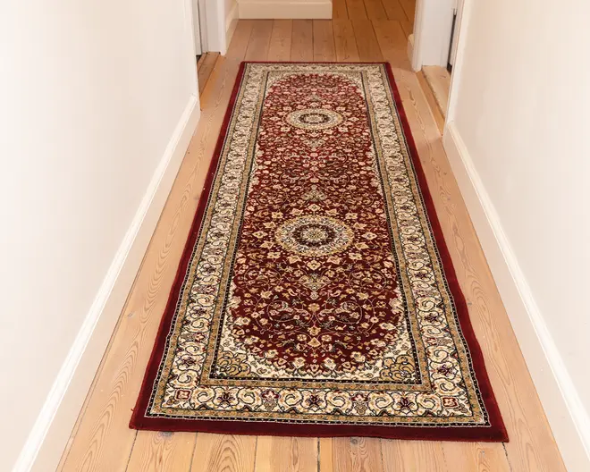 Lang, rektangulær tæppe med en dyb bordeaux baggrund og indviklede blomster-mønstre i creme, guld og sort. Tæppet har to store cirkulære medaljoner langs midten, omgivet af detaljerede vin- og blomstermotiver. En bred cremefarvet kant med dekorative mønstre omkranser tæppet, og kanterne er afsluttet med en ensfarvet bordeaux trim. Tæppet ligger på et lyst trægulv i en gang.