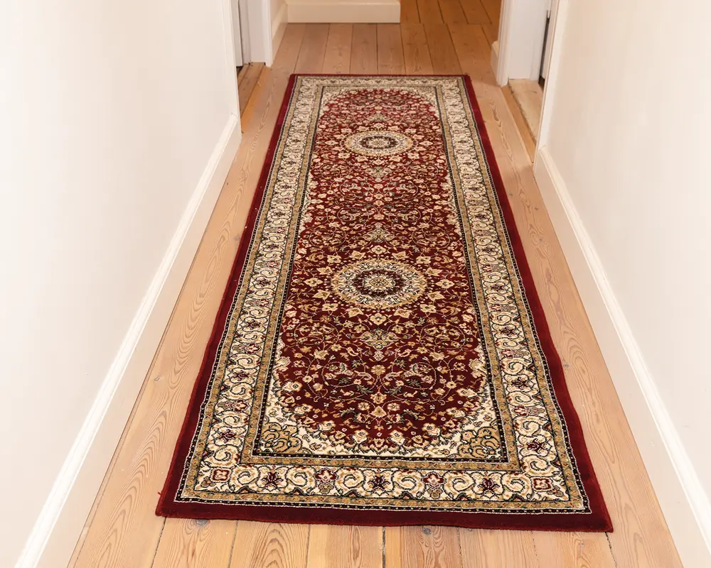 Lang, rektangulær tæppe med en dyb bordeaux baggrund og indviklede blomster-mønstre i creme, guld og sort. Tæppet har to store cirkulære medaljoner langs midten, omgivet af detaljerede vin- og blomstermotiver. En bred cremefarvet kant med dekorative mønstre omkranser tæppet, og kanterne er afsluttet med en ensfarvet bordeaux trim. Tæppet ligger på et lyst trægulv i en gang.