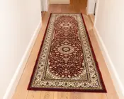 Lang, rektangulær tæppe med en dyb bordeaux baggrund og indviklede blomster-mønstre i creme, guld og sort. Tæppet har to store cirkulære medaljoner langs midten, omgivet af detaljerede vin- og blomstermotiver. En bred cremefarvet kant med dekorative mønstre omkranser tæppet, og kanterne er afsluttet med en ensfarvet bordeaux trim. Tæppet ligger på et lyst trægulv i en gang.