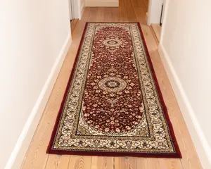 Lang, rektangulær tæppe med en dyb bordeaux baggrund og indviklede blomster-mønstre i creme, guld og sort. Tæppet har to store cirkulære medaljoner langs midten, omgivet af detaljerede vin- og blomstermotiver. En bred cremefarvet kant med dekorative mønstre omkranser tæppet, og kanterne er afsluttet med en ensfarvet bordeaux trim. Tæppet ligger på et lyst trægulv i en gang.