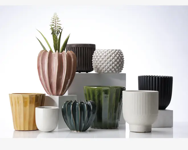 En samling keramiske urtepotter i forskellige former, størrelser og farver. Urtepotterne har forskellige overflader, herunder riflede, fløjlsbløde, facetterede og glatte finish. Farverne spænder fra pastelrosa, sennepsgul og mørkegrøn til hvid, sort og blå. Nogle potter har en blank glasur, mens andre har et mat udseende. Én potte indeholder en dekorativ plante med lange grønne blade og en hvid blomst.