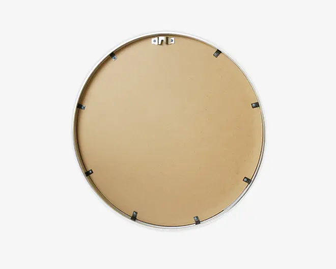 Billedet viser bagsiden af et rundt objekt med en glat, lysebrun overflade, sandsynligvis lavet af MDF eller lignende materiale. Øverst er der et metalophæng til vægmontering, og der er flere små sorte metalclips jævnt fordelt langs kanten, som holder bagbeklædningen fast. Yderringen ser ud til at være af metal eller malet sølv.