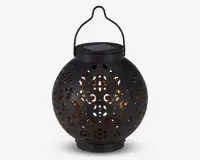 Rund sort lanterne med indviklede udskæringer i overfladen, som lader et varmt gult lys skinne igennem. Lanternen har et håndtag øverst til ophængning og et solcellepanel integreret i låget til opladning. Designet skaber dekorative lysmønstre, når den er tændt.