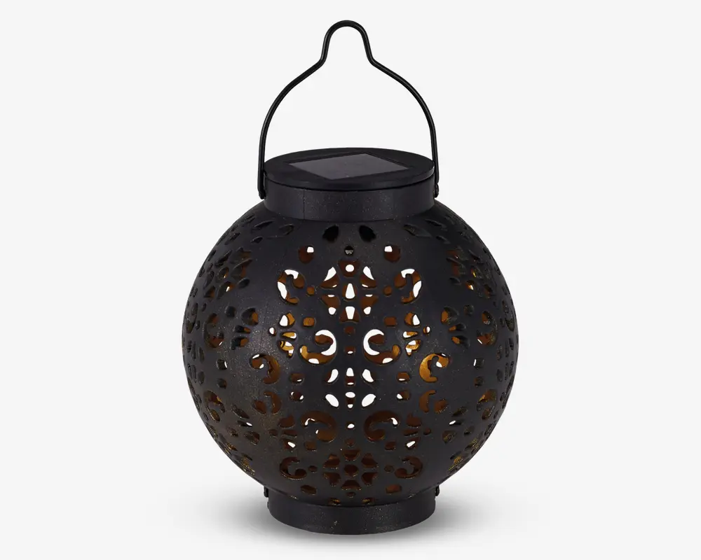Rund sort lanterne med indviklede udskæringer i overfladen, som lader et varmt gult lys skinne igennem. Lanternen har et håndtag øverst til ophængning og et solcellepanel integreret i låget til opladning. Designet skaber dekorative lysmønstre, når den er tændt.