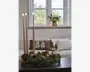 Rund grøn blomsterdekoration med tre høje, slanke stagelys, mørke bærdetaljer og en lav sort bakke som base.
