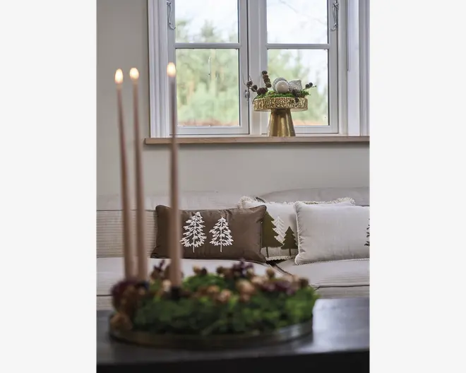 Rund grøn blomsterdekoration med tre høje, slanke stagelys, mørke bærdetaljer og en lav sort bakke som base.