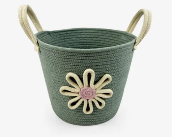 En rund, flettet kurv i en afdæmpet grøn farve med to solide, cremefarvede håndtag. På forsiden er der en dekorativ blomst lavet af cremefarvet snor med en lyserød midte, hvilket giver et legende og charmerende udtryk. Kurven har en tekstureret overflade og en bred åbning, velegnet til opbevaring af forskellige ting.