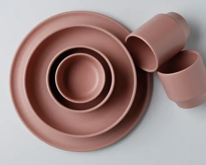Et sæt mat, rosafarvet keramikservice bestående af to tallerkener i forskellige størrelser, en lille skål og to kopper. Delene har et glat, minimalistisk design med blødt buede kanter og ensartet farve over hele overfladen.