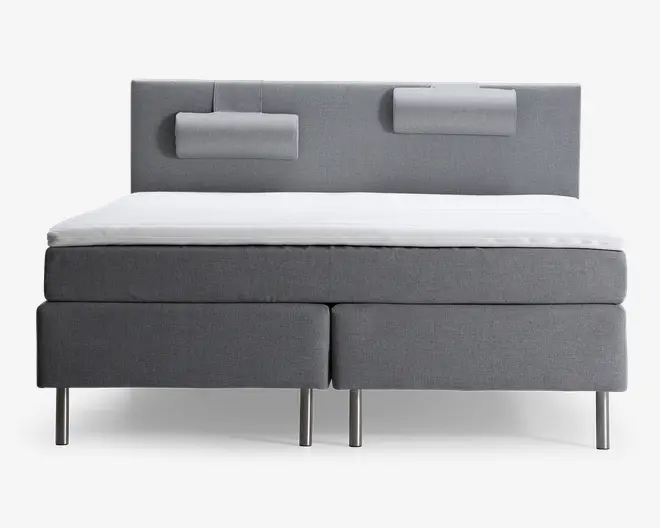 En moderne seng med gråt polstret stel og sengegavl. Sengegavlen har to rektangulære, polstrede puder fastgjort for ekstra komfort og støtte. Sengen har et rent, minimalistisk design med lige linjer og står på fire cylindriske metalben. Madrassen er dækket af en hvid topmadras, hvilket giver et frisk og indbydende udtryk.