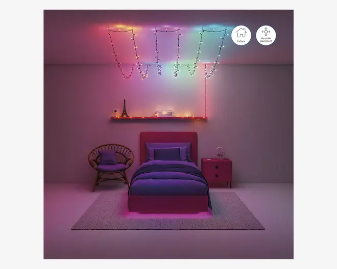 Et soveværelse med et moderne, minimalistisk design med en enkeltseng med polstret sengegavl og lilla sengetøj. Rummet er oplyst af flerfarvede lyskæder, der er arrangeret i geometriske mønstre i loftet og langs en svævende hylde over sengen. Hylden har dekorative genstande, herunder en lille Eiffeltårnsfigur og bøger. Til venstre for sengen står en rund kurvestol med en pude, og til højre et lille rødt natbord med en lampe. Gulvet er dækket af et lyst tæppe, og den samlede belysning skaber en hyggelig, farverig stemning.