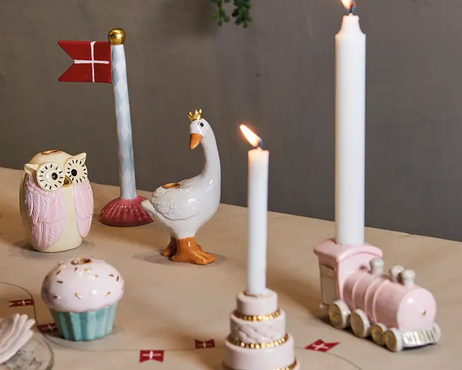 Billedet viser et dekorativt bordarrangement med flere keramiske figurer og lysestager. Der er en hvid gås med en guldkron, en lyserød og cremefarvet ugle, et lyserødt tog og en ornament formet som en cupcake. To hvide stearinlys står i pyntede holdere, hvoraf den ene har gulddetaljer. Der er også en stang med et rødt flag med hvidt kors. Genstandene er arrangeret på en lys dug, og små røde flag ligger spredt som ekstra pynt.